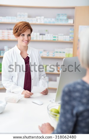 Attractive young female pharmacist standing holding a prescription smiling at the elderly patient, view over the shoulder of the patient