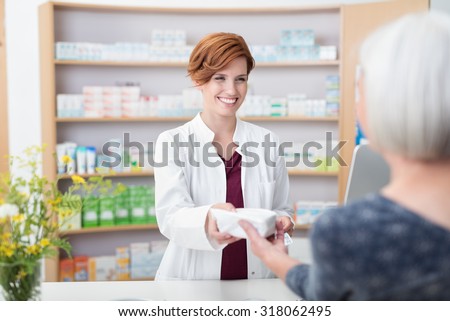 Smiling attractive young redhead pharmacist handing over prescribed medicines to an elderly female patient, view over the clients shoulder of the pharmacist