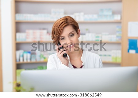 Pretty young female pharmacist talking on a telephone as she stands behind a computer in the pharmacy checking information with a smile