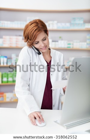 Young woman pharmacist talking on a phone as she stands behind her computer in the pharmacy checking information on a prescription she is holding