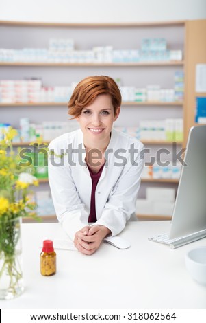 Friendly smiling young woman pharmacist leaning on the counter in the pharmacy with a bottle of pills in front of her smiling at the camera