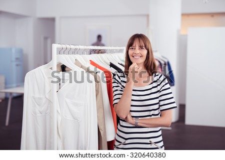 Half Body Shot of a Happy Adult Woman inside a Clothing Shop, Smiling at the Camera with One Hand on her Chin.