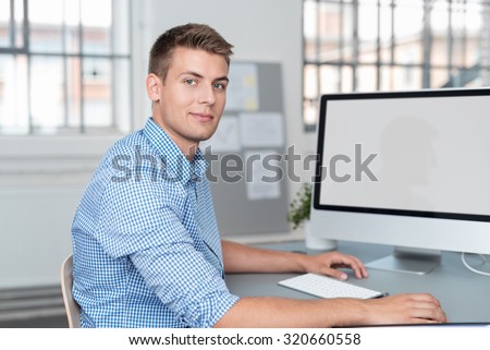 Handsome Young Businessman Sitting at his Desk with Blank Screen Computer, Smiling at the Camera.