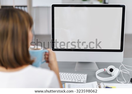 Businesswoman sitting back enjoying a mug of coffee and looking at a blank computer screen on a desktop computer which is visible to the viewer with white copyspace