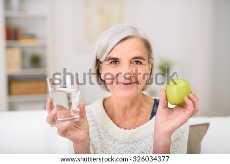 Portrait of a Gray Haired Senior Woman Holding Glass of Water and a Fresh Green Apple, Smiling at the Camera