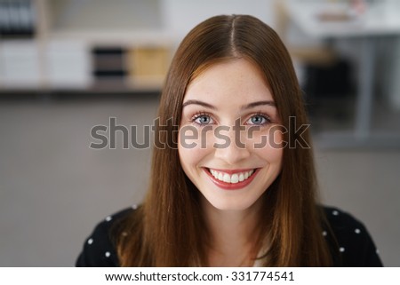 Gorgeous vivacious young woman with a beaming smile and happy eyes smiling directly at the camera, close up head and shoulders portrait indoors