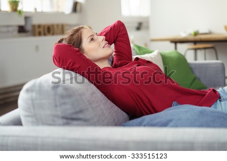 Young woman relaxing and daydreaming on a comfortable sofa at home staring up into the air with a smile and dreamy expression, side view