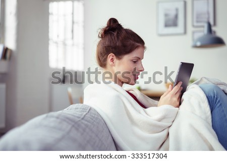 Happy young woman enjoying an e-book on her tablet computer as she relaxes with her feet up on the couch wrapped in a warm blanket, side view with copyspace