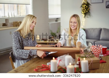 Two attractive young blond sisters smiling as they wrap Christmas gifts together seated at the dining table in their living room at home