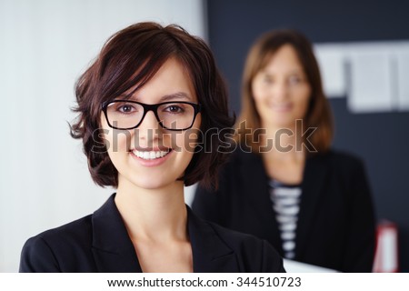 Smiling attractive manageress or team leader posing in the foreground with a team member or co-worker visible in the background as a blur, head and shoulders portrait