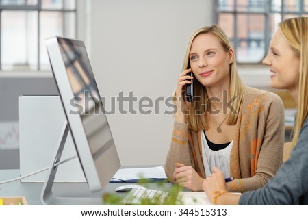 Pretty Office Woman Talking to Someone Using Phone While Working on the Computer with her Female Colleague.