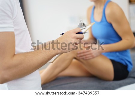 Male Physical Therapist, Holding Pen and Paper, Interviewing his Female Patient with Knee Injury, Sitting on the Bed Inside the Clinic.