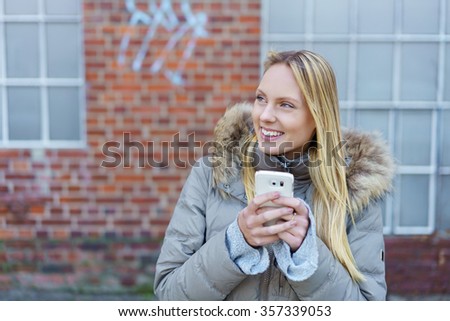 smiling blond woman with cellphone standing in front of a brick wall on the street