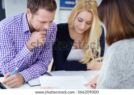 Broker giving a presentation to a young couple in her office leaning over the desk to explain paperwork to them