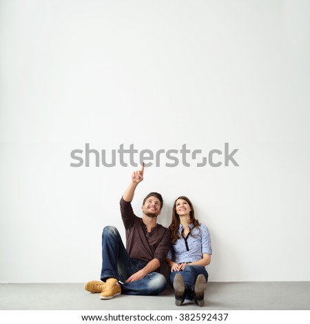 Young couple sitting on the ground leaning against an exterior white all with copy space pointing up into the air and smiling