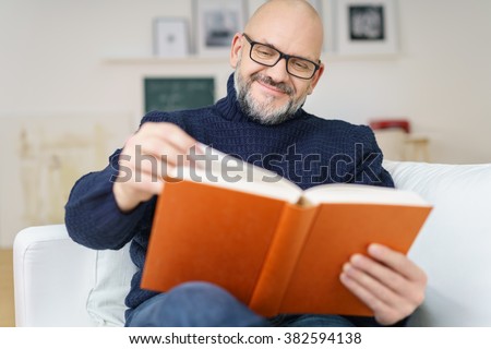 Middle-aged bald man with a goatee wearing glasses sitting on a comfortable couch enjoying a good book with a smile of pleasure
