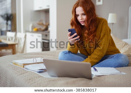 Attractive Young Woman with Red Hair Working from Home - Female Entrepreneur Sitting on Bed with Laptop Computer, Paperwork and Checking Cell Phone from Comfort of Home