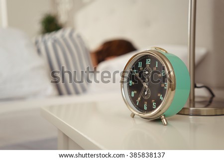Alarm clock about to ring alongside a sleeping person in bed with focus to the bedside table ad clock