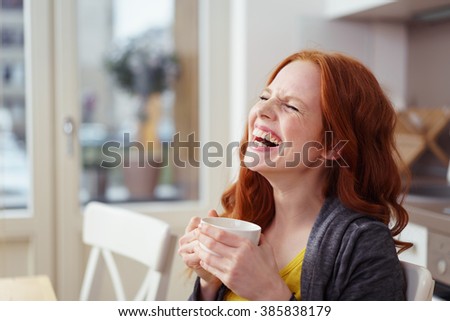 Spontaneous attractive young redhead woman enjoying a good laugh over a morning cup of coffee at home in the apartment