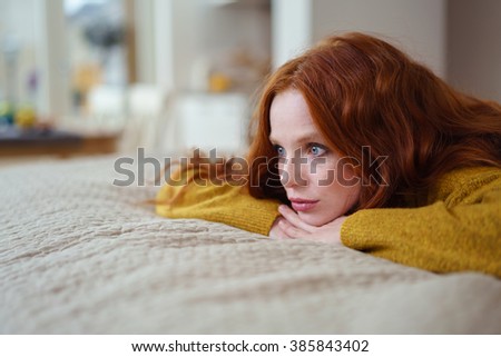 Attractive young redhead woman lying on her bed on her stomach thinking resting her chin on her hands and staring into the distance