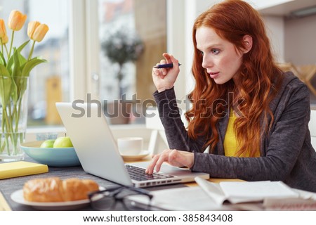 Serious beautiful young female in gray sweater and long red hair seated at table as she types on her laptop next to papers, eyeglasses and croissant