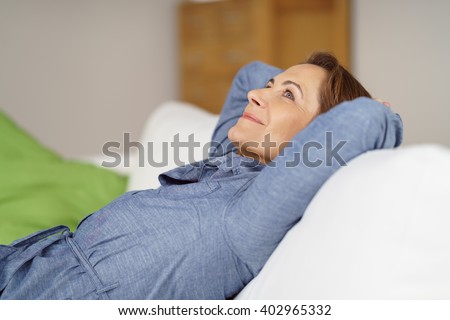 Happy middle aged woman relaxing at home reclining on a comfortable sofa with her hands behind her head looking upwards with a dreamy smile of pleasure