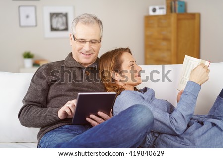 Happy middle-aged married couple relaxing together at home with the wife lying back against her husband as she reads a book while he surfs the internet on a tablet