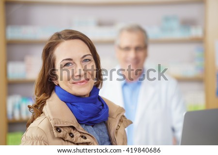 Attractive stylish middle-aged woman with a lovely smile standing looking at the camera in a pharmacy with a male pharmacist visible as a blur behind