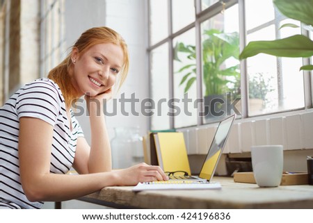 Smiling happy young woman studying at home sitting at a work table with her notes and laptop looking at the camera with a friendly grin