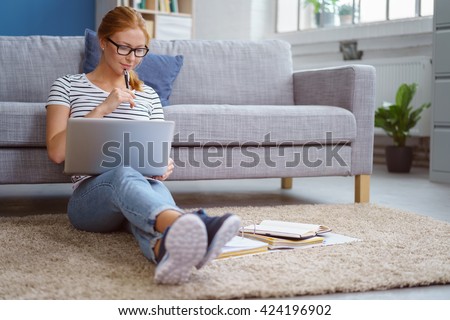 Studious young woman working at home sitting on the floor in the living room with a laptop and class notes in binders studying for university