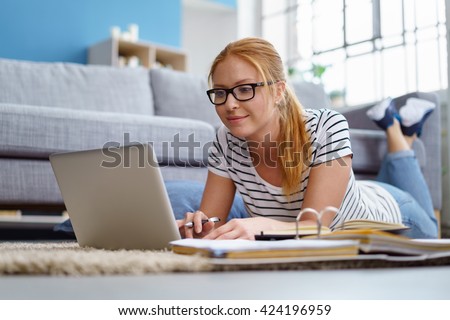 Smiling young female lying on the floor in the living room student studying at home working with her laptop computer and class notes