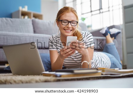 Happy young woman studying at home reading an sms or text message on her mobile phone with a smile as she lies on the floor with a laptop
