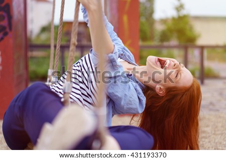 Carefree pretty young redhead woman frolicking in an urban park playing on a rope swing and enjoying a hearty laugh
