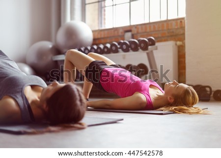 Two young adult women doing pelvic muscle exercises on individual rectangular mats with weights and stability ball in background at gym