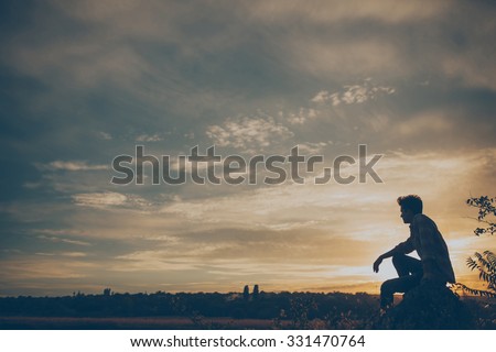 Silhouette of young man sitting on sunset or sunrise. Confident teenage boy thinking on cliff stone. Hope. Sadness. Freedom.