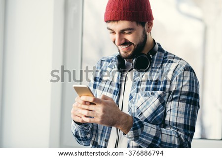 Handsome happy hipster young man using smart phone at window. Male wearing headphones and shuffling music