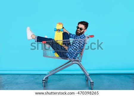 Carefree hipster fun. Side view of cheerful young man sitting in shopping cart with bright skateboard over colorful wall