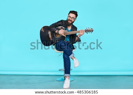 handsome bearded musician playing guitar over blue background. Full length man sitting on chair holding acoustic guitar