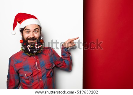 Happy hipster man in santa hat with funny christmas decorated beard pointing to the side at copy space over white and red background