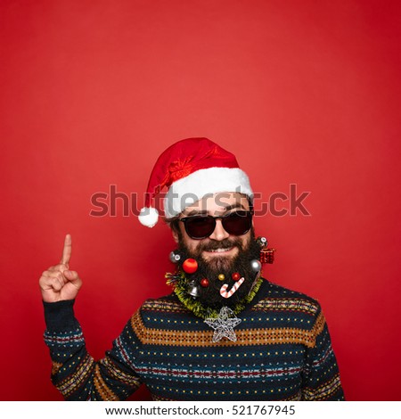 handsome man with decorated christmas beard and moustache in red santa claus hat gesturing up with his finger to copy space above his head