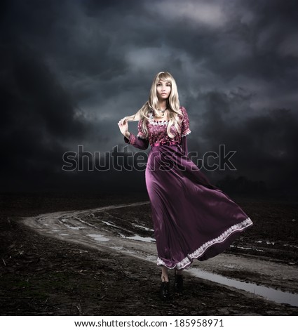 Full Length Portrait of Fantasy Woman in Waving Purple Dress Walking on Dirty Road. Moody Sky. HDR Cloudscape.