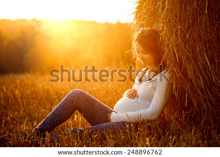 Young Pregnant Woman Sitting by the Haystack at Sunset and Embracing her Belly. 7 Month Pregnancy. Maternity Concept. Toned Photo.