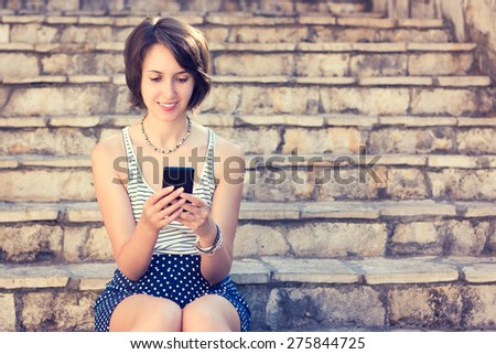 Young Hipster Girl Texting with Her Mobile Phone and Smiling Outdoors. Modern Youth Lifestyle, Technology and Communication Concept. Toned Photo.