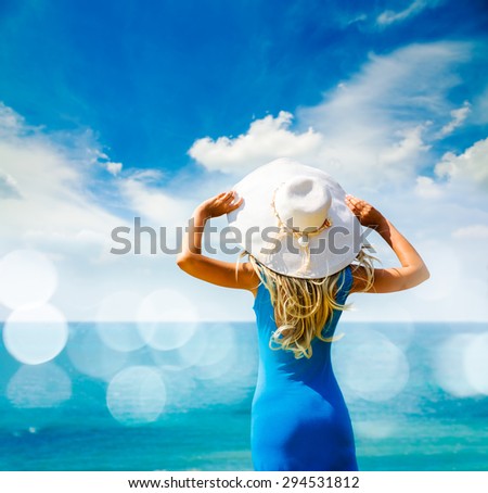 Blonde Woman in Blue Dress Standing at Sea and Holding White Hat. Back View. Summer Vacation Concept. Photo with Bokeh and Copy Space.