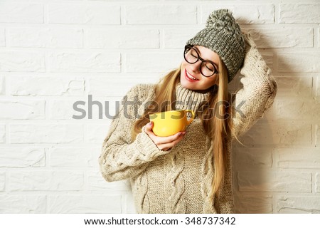 Romantic Dreaming Hipster Girl in Knitted Sweater and Beanie Hat with a Mug in Hands at White Brick Wall Background. Winter Warming Up Concept.