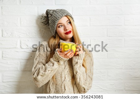 Romantic Dreaming Winter Hipster Girl in Knitted Sweater and Beanie Hat with a Mug in Hands at White Brick Wall Background. Warming Up Concept.