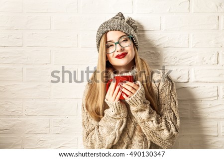 Romantic Winter Hipster Girl in Knitted Sweater and Beanie Hat Enjoying a Cup of Hot Tea in Hands. Lovely Dreaming Woman. White Brick Wall Background. Warming Up Concept. Toned Photo with Copy Space.