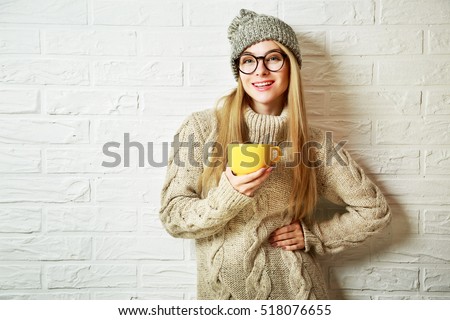 Happy Fashion Hipster Girl in Knitted Sweater and Beanie Hat with a Mug in Hands. Smiling Nice Woman at White Brick Wall Background. Winter or Autumn Warming Up Concept. Toned Photo with Copy Space.