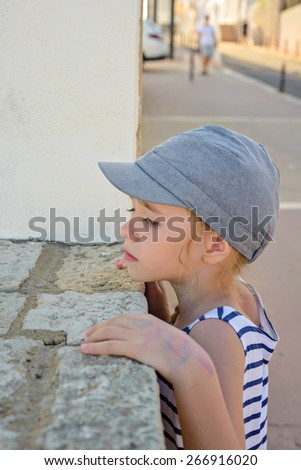 Little girl in denim cap and striped clothes looking through a wall in the city