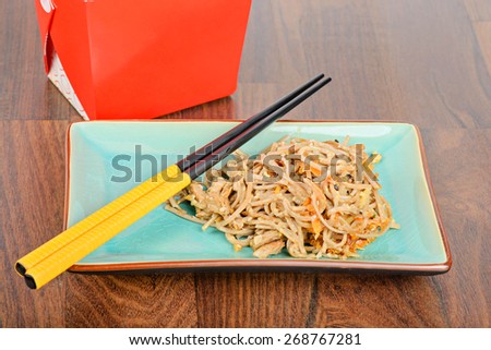 Meat and noodles in blue plate with chopsticks on wooden table. Red take away box on the background.